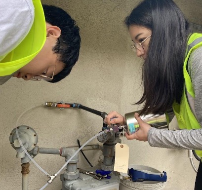 Two researchers in safety vests collecting a natural gas sample from a residential outdoor gas meter into a stainless steel sample canister.